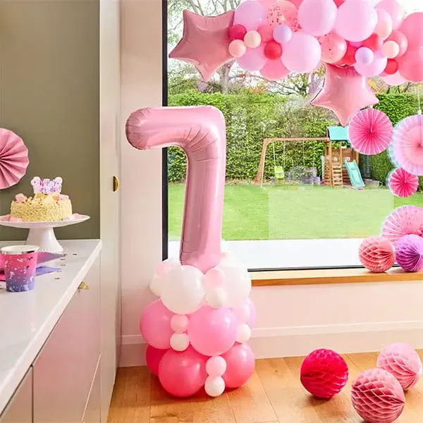 Pink number 7 balloon display with a pink balloon arch, paper decorations, and birthday party décor beside a window.